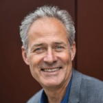 Lächelnder Mann mittleren Alters mit grauem Haar, bekleidet mit einem blauen Hemd und einem grauen Blazer, steht vor einem schlichten braunen Hintergrund.