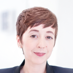 Eine Person mit kurzem, hellbraunem Haar und heller Haut, die einen dunklen Blazer trägt, lächelt sanft in einer hellen, verschwommenen Innenumgebung.