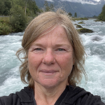 Eine Frau mittleren Alters mit blondem Haar macht ein Selfie vor einem fließenden Fluss, umgeben von grünen Bäumen und Bergen unter einem bewölkten Himmel.