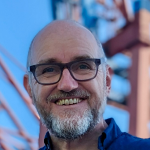 Ein lächelnder Mann mittleren Alters mit Brille, Glatze und grauem Bart steht im Freien vor einer verschwommenen Industrieanlage.