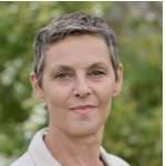 Eine Person mit kurzen grauen Haaren und heller Haut steht im Freien und blickt mit neutralem Gesichtsausdruck in die Kamera. Der Hintergrund ist grün und unscharf und lässt an Laub oder Bäume denken.