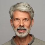 Ein lächelnder älterer Mann mit grauem Haar und Bart, der ein graues Hemd mit Kragen trägt und vor einem schlichten hellgrauen Hintergrund posiert.