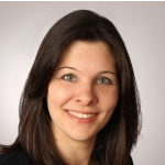 Eine Frau mit glattem braunem Haar und blauen Augen lächelt in die Kamera. Sie trägt ein schwarzes Oberteil und posiert vor einem schlichten, hellen Hintergrund.